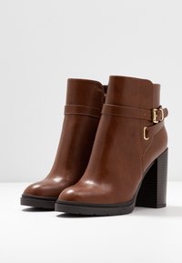 Hnědé kožené kotníkové boty s masivním podpatkem a přezkou. Hladká textura, kulatá špička a kontrastní černá podrážka.