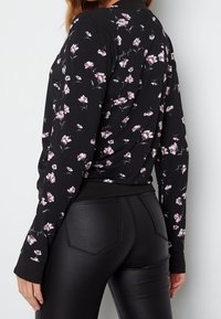 Femme portant un haut florale noir à manches longues et un pantalon en simili cuir noir, vue de dos contre un fond clair uni.