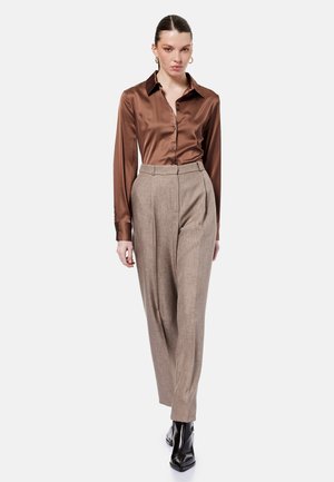 Mujer caminando con una camisa marrón de satén abotonada, pantalones beige de talle alto, botas negras puntiagudas y pendientes de aro dorados sobre fondo blanco.