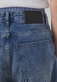 Modré džínové kraťasy s koženou nášivkou s raženým textem, viditelnými zadními kapsami a texturovaným pasem s vertikálními pruhy.