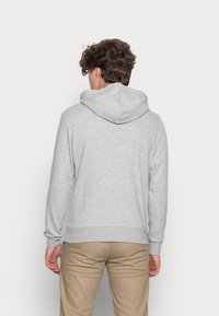 Hombre con cabello rizado que lleva una sudadera con capucha gris claro y pantalones beige, de pie frente a un fondo blanco liso.