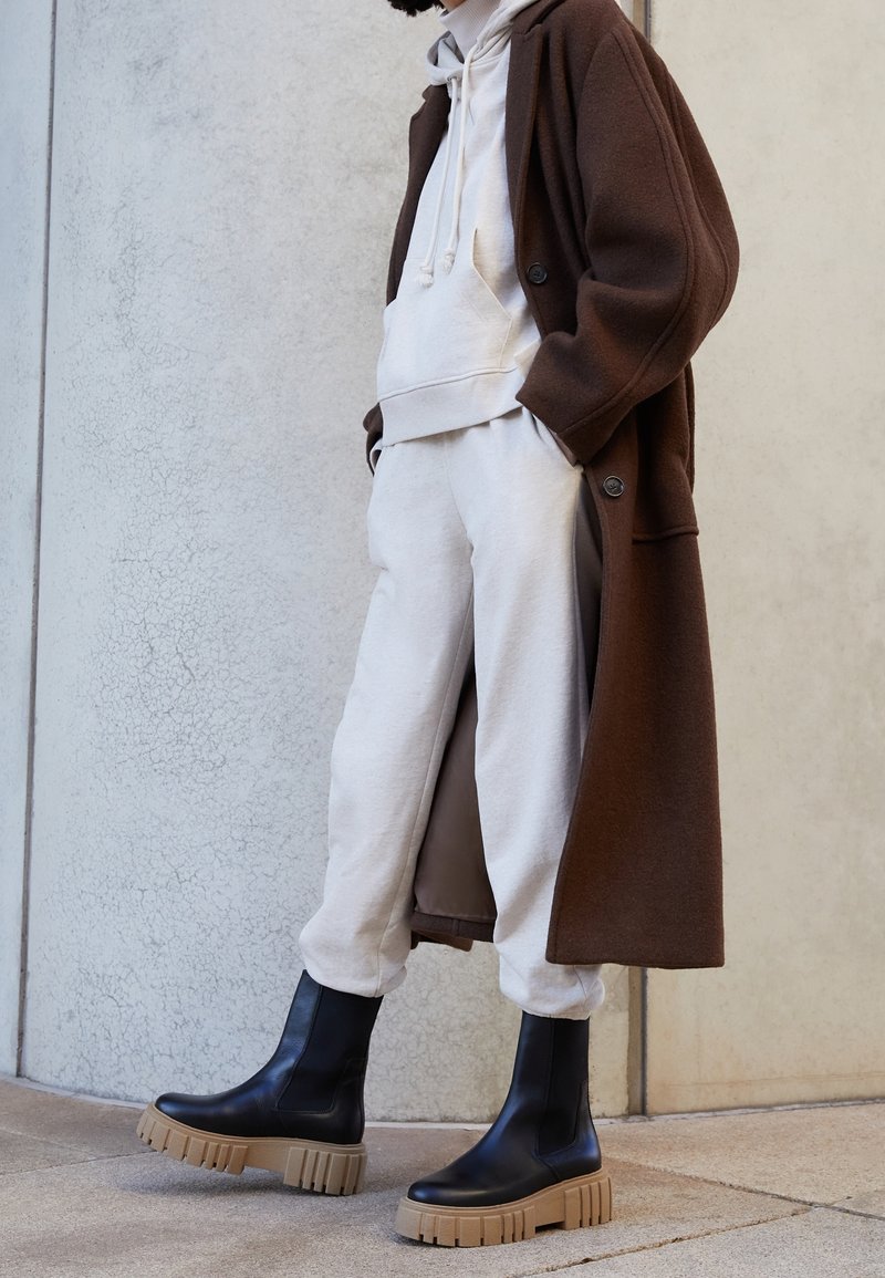 Personne portant un sweat à capuche blanc et un pantalon de survêtement assortis à des bottes noires épaisses et un long manteau marron debout contre un mur texturé.