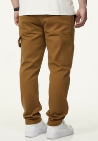 Brown cargo pants with side pockets, tapered fit, and a smooth texture, paired with white sneakers. Back view showcases stitching details.