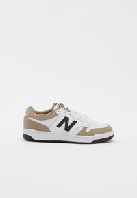 Valkoiset ja beige New Balance -tennarit mustalla "N"-logolla, valkoiset nauhat ja rei'itetty kärki valkoista taustaa vasten.