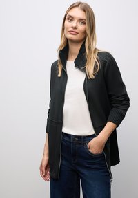 Giacca nera con zip e colletto a coste, tasche laterali e polsini a coste, abbinata a una t-shirt bianca e jeans blu scuro.