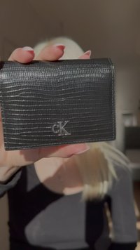 Zwarte leren portemonnee met een getextureerd, krokodillenachtig patroon; heeft een zilveren "CK" logo aan de voorkant. Compact ontwerp, rechthoekige vorm.