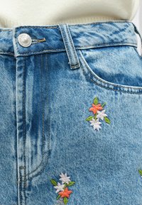 Falda vaquera en azul claro con bordados florales en naranja, blanco y verde. Cuenta con un cierre frontal de botones y un diseño clásico de cinco bolsillos.