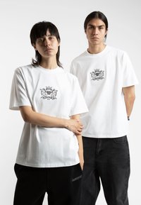 White cotton t-shirts with short sleeves, featuring a black logo design on the chest and back. Loose fit, paired with black pants.