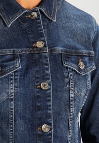 Blå denimjacka med metal snapknappar, en spetskrage och två bröstfickor med lock. Tyget visar typisk denimtextur.