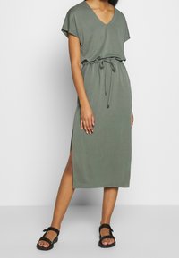 Mujer vistiendo un vestido midi verde oliva con escote en V, cintura con cordón, abertura en el costado y sandalias de tiras negras, de pie sobre un fondo plano.