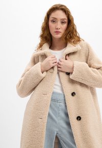 Beige Fleece-Mantel mit Kragen, ausgestattet mit schwarzen Knöpfen und einer strukturierten Oberfläche. Getragen über einem weißen Oberteil und hellblauen Jeans.