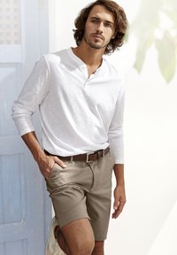 Maglietta henley bianca a maniche lunghe abbinata a pantaloni corti beige, con una cintura marrone. Sullo sfondo si intravede una porta azzurro chiaro. Abbigliamento estivo informale.