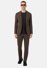 Ruskea pikkutakki, jossa on muotoiltu fit, yhdistettynä tummansiniseen t-paitaan ja sopiviin ruskeisiin housuihin. Mustat kengät viimeistelevät asun.