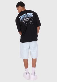 Black oversized t-shirt with graphic text on the back, paired with light denim shorts, white high-top sneakers, and white socks.