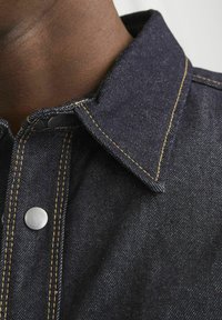 Camicia in denim scuro con colletto a punta, dotata di bottoni a pressione argentati, cuciture gialle in contrasto e texture liscia.