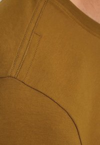 Horalová tričko v horčicovej farbe s hladkým materiálom, okrúhlym výstrihom a jemnými švami blízko ramena a lemu.