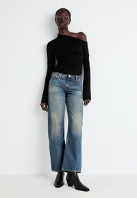 Top negro de manga larga y escote off-shoulder con textura fruncida, combinado con pantalones vaqueros de pierna ancha en azul claro con efecto desteñido. Botines negros de tacón.