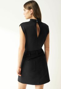 Zwarte jurk met korte mouwen, een sleutelgatopening aan de achterkant en drie knopen bij de hals. Beschikt over twee grote achterzakken en een getailleerde taille.