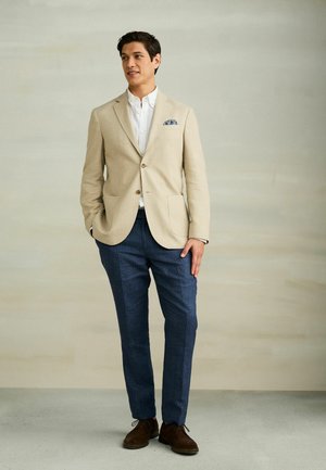 Mężczyzna w beżowej marynarce, białej koszuli, niebieskich spodniach i brązowych butach, stoi z jedną ręką w kieszeni na neutralnym tle.