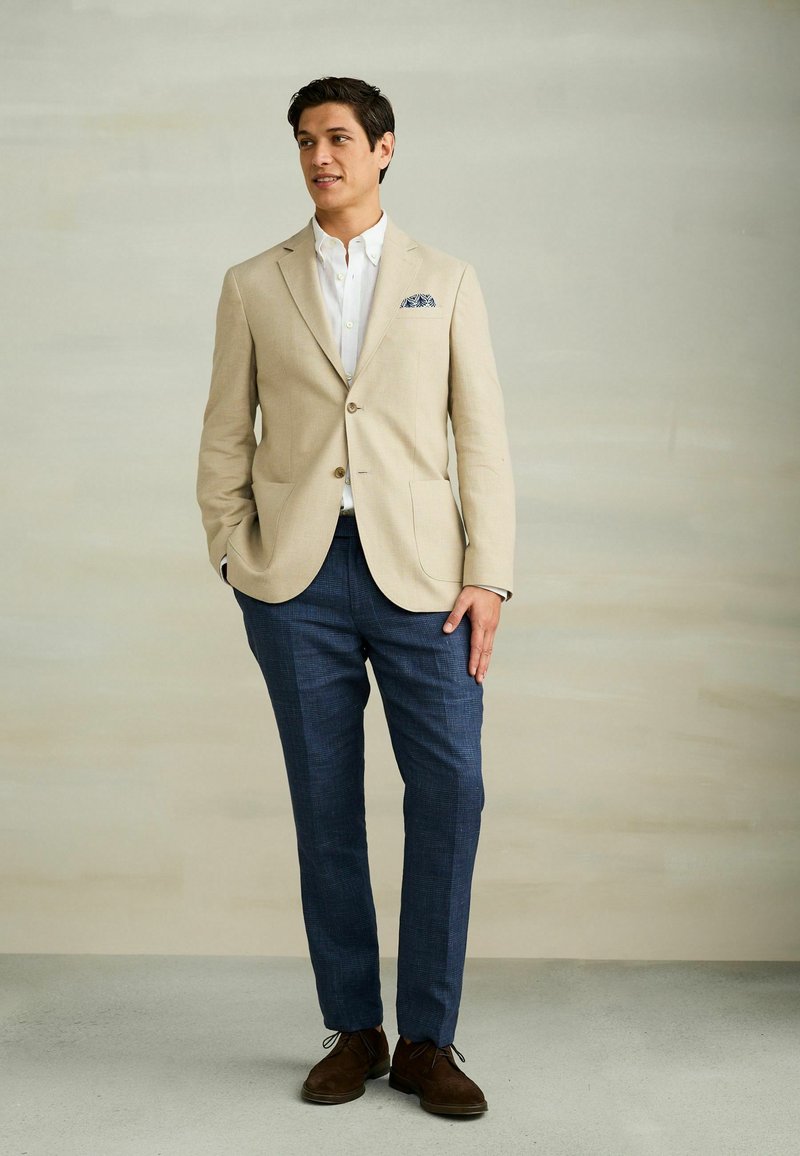 Homme portant un blazer beige, une chemise blanche, un pantalon bleu et des chaussures marron, debout avec une main dans la poche sur un fond neutre.