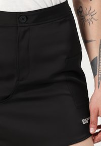 Crne kratke hlače od glatkog, rastezljivog materijala s prednjim kopčanjem na gumb, bočnim džepovima i detaljem s logom na rubu.