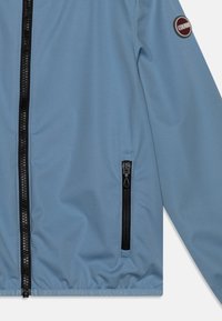 Niebieska kurtka softshell z czarnym zapięciem na zamek, detalem w postaci bocznych kieszeni oraz okrągłą łatą z logo na lewym rękawie. Gładka faktura.