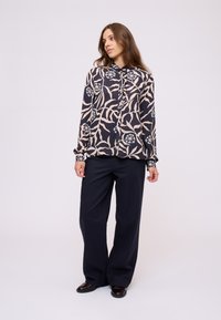 Donna con lunghi capelli castani che indossa pantaloni neri a gamba larga e una blusa floreale scura con motivi di foglie rosa chiaro e blu, in piedi su uno sfondo neutro.