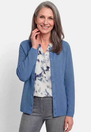 Glurende middelbare vrouw met grijs haar draagt een blauw gebreid vest, een blouse met patroon en grijze broek, staande tegen een effen achtergrond.