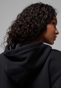 Mujer con cabello rizado y oscuro que lleva una prenda con capucha negra, mostrada de perfil contra un fondo gris liso.