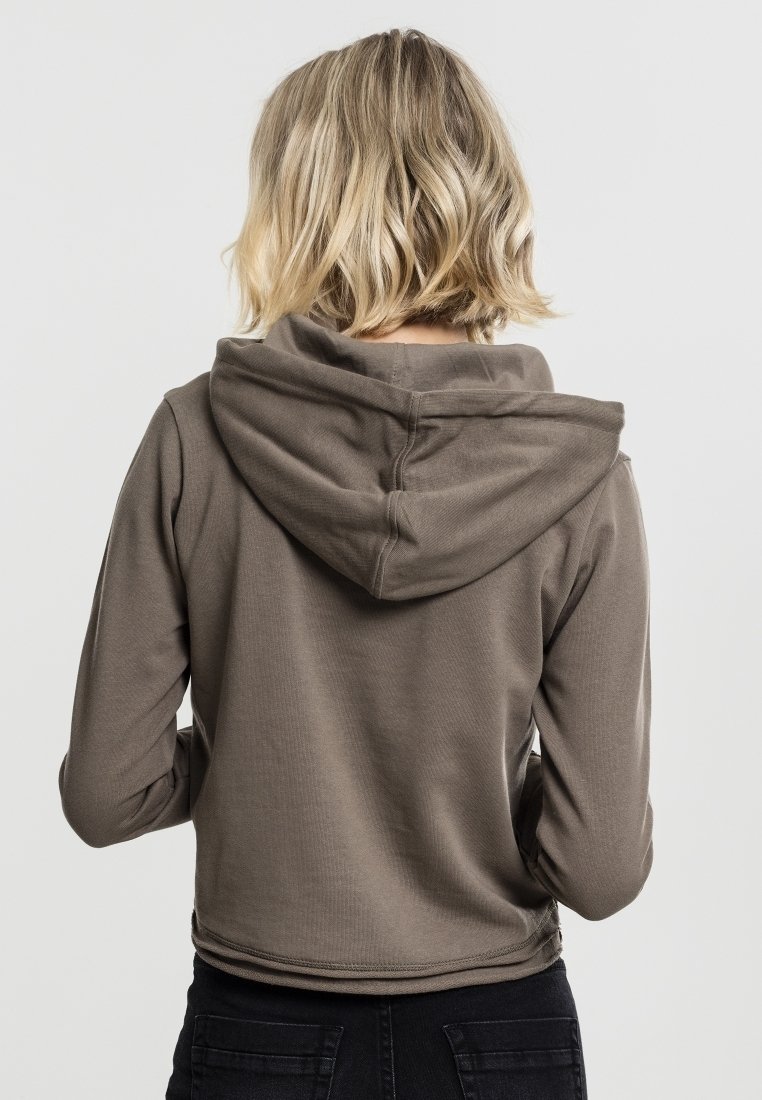 Personne blonde portant un sweat à capuche marron et un pantalon noir, vue de dos sur un fond clair uni.