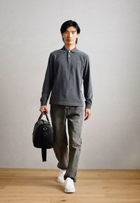 Γκρίζο μακρυμάνικο πόλο μπλουζάκι, ανοιχτό γκρι τζιν, λευκά αθλητικά παπούτσια, κρατώντας μια μαύρη δερμάτινη τσάντα ταξιδιού με γυαλιστερό ασημί φερμουάρ και χερούλι.