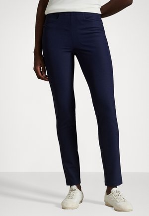 Pantaloni slim-fit blu navy. Presentano una superficie liscia, tasche frontali e cuciture sottili. Abbinati a sneaker bianche.