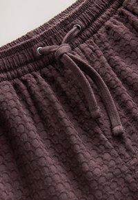 Brązowe szorty o teksturowanej powierzchni z wzorem plastra miodu, elastycznym pasem i sznurkiem do regulacji. Materiał wydaje się miękki i wygodny.