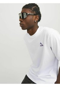 T-shirt bianca a maniche corte con un logo triangolare viola sulla parte sinistra del petto, abbinata a grandi occhiali da sole neri. Tessuto dalla texture liscia.