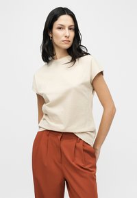 Μπεζ βαμβακερό t-shirt με κοντά, γυρισμένα μανίκια και άνετη εφαρμογή. Συνδυασμένο με ψηλόμεσα, πιεττέ παντελόνια σε σκουρόχρωμη σκουριά.