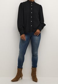 Blouse noire à manches longues avec un col montant, ornée de boutons en perles et d'une dentelle sur le devant, assortie à un jean skinny bleu et des bottines brunes.