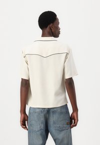 Short-sleeve cream shirt with dark piping, a curved yoke at the back, and a relaxed fit, paired with light blue jeans.