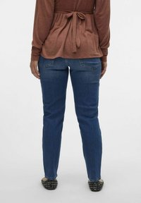 Hnědý svetr s dlouhým rukávem a detailem na vázání v pase, spárovaný s modrými džíny. Džíny mají klasický střih a zadní kapsy; nošeny s barevnými balerínkami.