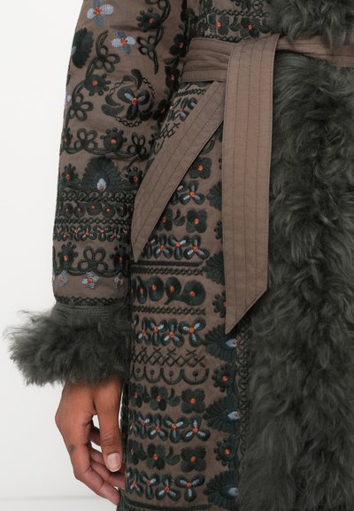 Καφέ κεντητό παλτό με πράσινα λουλουδάτα μοτίβα, που διαθέτει μπορντούρα από γούνα και ζώνη. Υφή υφάσματος με περίπλοκες ραφές και λεπτομέρειες.