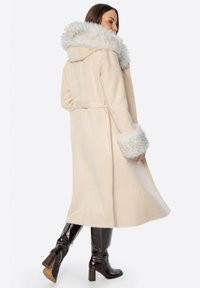 Kremowy długi płaszcz z futrzanym kapturem i mankietami. Posiada pasek w talii oraz teksturowany materiał. Gładki, pełnej długości fason.