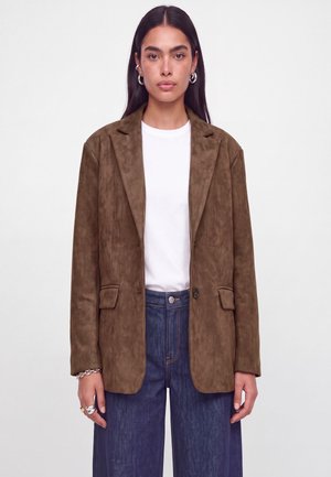 Femme portant un blazer en daim marron sur une chemise blanche et un jean bleu, debout sur un fond blanc uni.