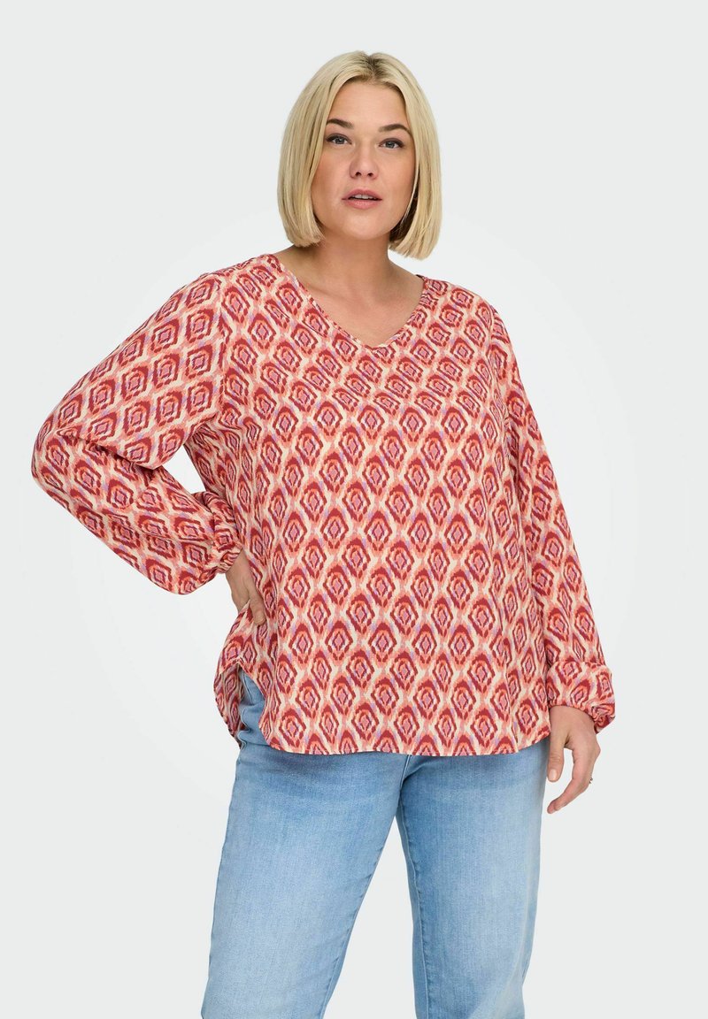 Červená vzorovaná halenka s výstřihem do V a dlouhými rukávy, s diamantovým vzorem v odstínech červené a krémové, v kombinaci se světle modrými džínami.