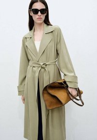 Světlý olivově zelený trench coat s páskem, širokými klopami, dlouhými rukávy a hladkým materiálem. Modelka drží hnědou semišovou tašku s koženými akcenty.