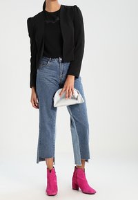 Mujer con un blazer negro corto y top, jeans cortos bicolor, botas de tobillo rosas brillantes, sosteniendo un pequeño bolso de mano plateado.