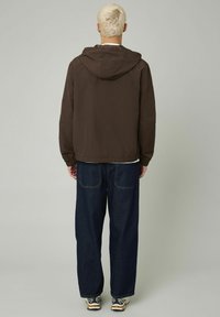 Veste à capuche légère marron, coupe décontractée, vue de dos, associée à un jean large bleu foncé et des baskets beiges.