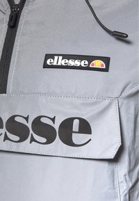 Gray windbreaker featuring a large black "ellesse" logo on the pocket and a smaller rectangular brand patch near the zipper. Smooth texture.