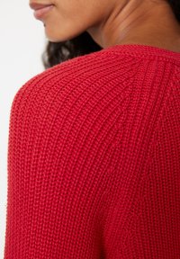 Czerwony sweter z dzianiny, charakteryzujący się teksturowanym wzorem prążków, szerokim dekoltem oraz lekko luźnym krojem, podkreślający miękką tkaninę i detale wzornicze.