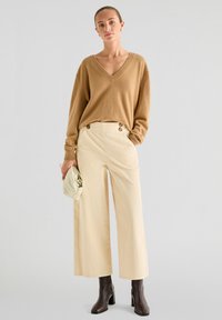 Brun strikket sweater med V-hals, cremefarvede vide corduroy bukser med knapdetaljer og brune hælesko; holder en lys taske.
