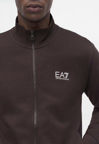 Homme portant une veste zippée marron foncé avec un col montant et le logo blanc EA7 Emporio Armani sur le côté gauche de la poitrine.