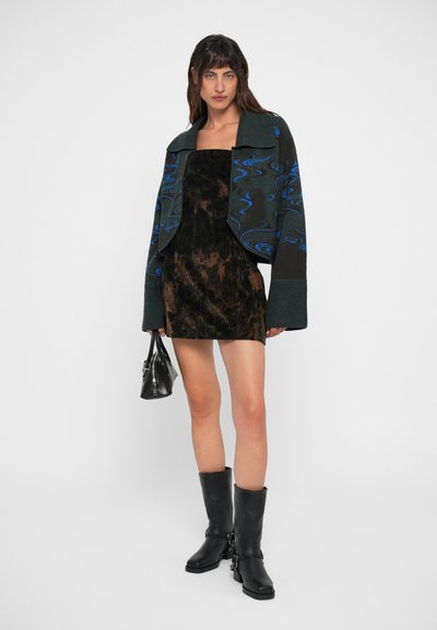 Vestido mini de veludo preto com uma jaqueta cropped texturizada e estampada em verde e azul. Modelo usa botas altas pretas e segura uma pequena carteira.